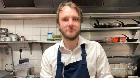 A chef wearing a navy apron with his sleeves rolled up is standing in a restaurant kitchen. There are pots and pans in the background.