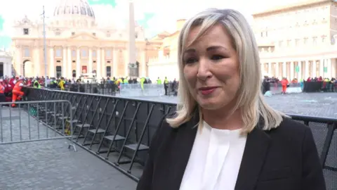 Michelle O'Neill, dressed formally in black with a white blouse, smiling as she speaks to a journalist.
