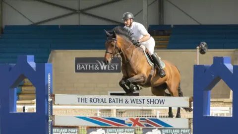 Gorman Photography A showjumper rides his horse as it leaps over a barrier in the Main Ring. "The Royal Bath and West Show" is written on the barrier and a further sign reading "Bath and West" can be seen on a wall in the background.