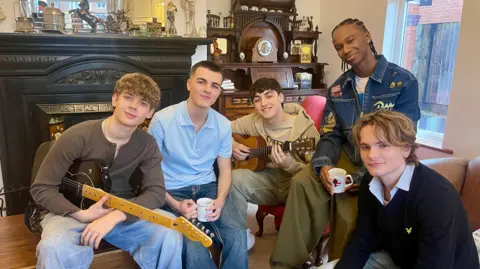 Five young men sit in a dated living room, two of whom are holding guitars and two hold mugs of tea. The man on the left is holding a black electric guitar, has short curly hair and is wearing a brown long-sleeved top and blue jeans. Next to him is a man in a blue polo shirt and blue jeans holding a white mug. A man with black hair, blue jeans, sand hoodie and sand t-shirt is playing a brown acoustic guitar. To the right of him, sat slightly higher on the arm of a brown settee, is a man holding a white mug with black cornrows, a white t-shirt, blue denim jacket and green trousers. Sat on the settee to his right is a man with long fair hair, a blue shirt and a black jumper. Behind the men is a large black fireplace covered in ornaments in front of a mirror. There is also a brown antique desk with a clock next to a window.