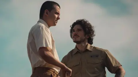 Asa Butterfield standing beside Fra Fee. Butterfield is wearing a short sleeve, cream coloured shirt. He has short brown hair. Fee is wearing a short sleeve, green shirt. He has short, curly brown hair. 