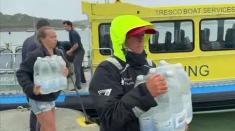Two people collect bottled water from a bottled water station set up on Bryher's quayside. One of the people is wearing a raincoat with a luminous yellow hood. A yellow boat is in the background.