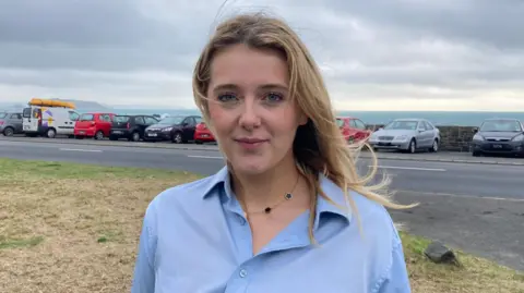 BBC A woman standing outside with a row of cars parked in front of the ocean, a road in between and grasslands directly behind her. She is wearing a blue shirt and the sky is grey and cloudy.