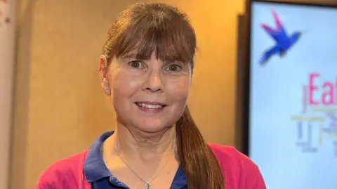 Karen is wearing a navy polo shirt and pink cardigan stands indoors beside a tall banner displaying the JEDS logo with a stylized bird graphic. A presentation screen with a word cloud is visible behind them.