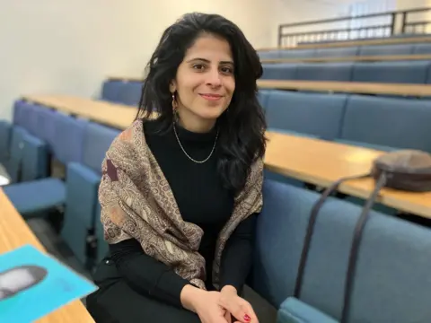 Lina al-Hathloul. She has long black hair and has a shawl around her shoulders. She has a gold necklace and sits on blue fold-down in what appears to be a meeting room.