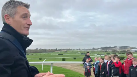 Jockey Daryl Jacobs is standing in front of a group of school children on the track at Cheltenham Racecourse. He is wearing a navy coat. The children are stood opposite him and listening to his stories. Cheltenham Racecourse grandstand is in the distance in the background. 