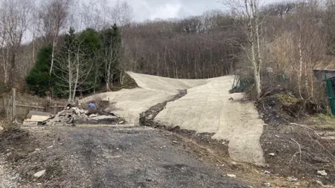 The landslide scene, resembling a dry ski slope, with protective matting laid over the bare earth to prevent further erosion.  A demolished garage can be seen to the side, which was flattened by the landslide.  There are trees in the background and the ground is black.  