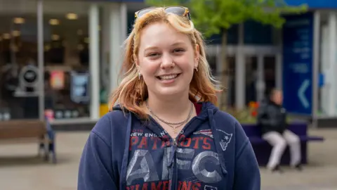 Close up image of Alex smiling. She is stood in Bangor town centre. She wears a navy zip up hoodie with red writing on, silver necklaces and has ginger wavy hair. 