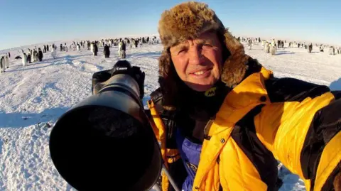 Doug Allan - selfie of man in puffer jacket and woolly hat, holding large camera in a snowy location surrounded by penguins
