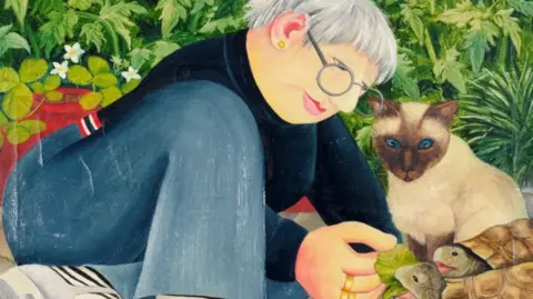 ourberylcook.com/John Cook A woman in a blue top and navy jeans is sitting outside feeding two blue-shelled tortoises with lettuce whilst a cream Siamese cat with a brown face and blue eyes watches.