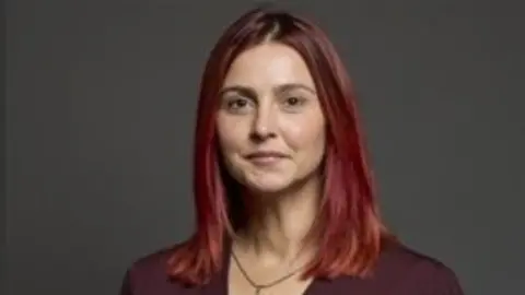 Melanie Onn has shoulder length red hair and she is wearing a plum top with a gold necklace. The image is a head and shoulders portrait. 