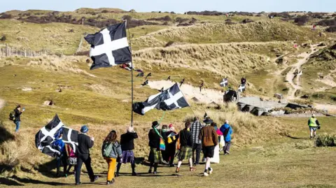 Kai Greet photography A group of people walking towards grassy dunes. several of them are holding flags. 