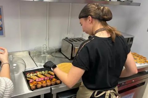 A person in an apron and hair net working to garnish some canapes.
