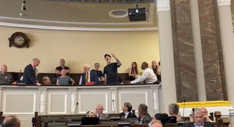 A video still of members of the public sat in a gallery above councillors in the city council chamber. One man, dressed in a black T-shirt and a black hat, is stood up and gesturing with a raised arm towards the councillors.