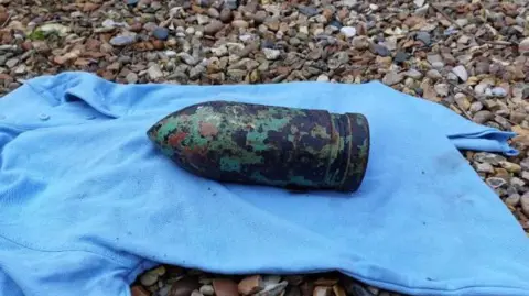 A rusted "unexploded device" in the shape of a large bullet-shaped shell, is on the ground on top of a light blue T-shirt. 