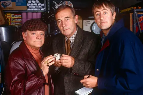 A television promotional picture showing three men standing shoulder to shoulder as they hold a small pocket watch with a white face. The man on the left is wearing a checked flat cap, a burgundy leather jacket and an orange polo-neck sweater. The man in the midden is wearing a tweed jacket, checked shirt and brown tie. The man on the right is younger and is wearing a blue coat and holding a notepad and pen.