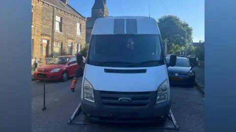 Front view of a white Ford Transit-style van parked on a residential street and positioned on the platform of a recovery truck. Brick terraced houses line the street on the left, with several parked cars visible on both sides, including a red car and a dark blue car. A church tower rises in the background behind the van. Bright sunlight casts strong shadows, and part of a person wearing high-visibility orange trousers is visible beside the recovery vehicle. The van's windscreen is darkly tinted, and patterned