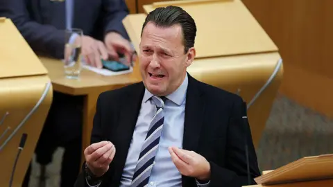 Getty Images Russell Findlay has slicked-back brown hair and is wearing a black jacket, grey shirt and grey and black striped tie
