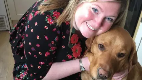 Family photograph Gemma Fairhurst, smiling with blonde hair, wears a black dress with a flowery pattern. She is crouched down with her face cheek-to-cheek with guide dog Ian.