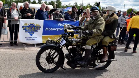 VMCC A man in old-school motorcycle clothing sits on a vintage bike with a woman on the back. They are both wearing helmets. Crowds of people are watching from the other side of a barrier.