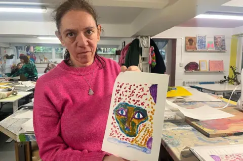 BBC / Rosa Rahimi A woman looking at the camera and holding up a painting on an A4 piece of paper. The painting shows a yellow, green and blue face surrounded by yellow and purple dots. She is wearing a pink jumper, and is stood in an art studio.