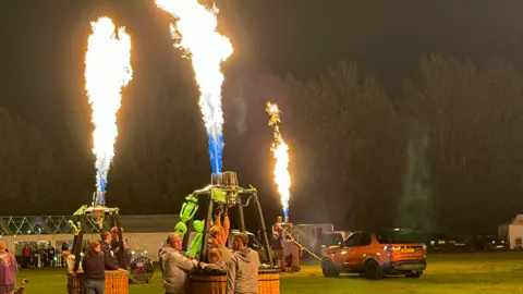 Hot air balloons firing their burners while on the ground, showing flames leaping into the night sky