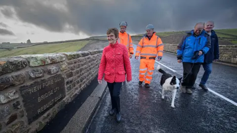 North Yorkshire Council The image shows a group of five people walking across a stone bridge.  The bridge has a plaque on the left-hand side that reads:
“NEW FORE GILL BRIDGE 271 REBUILT 2025.”
Two of the people are wearing bright orange high-visibility workwear, one is in a red quilted jacket, and another two are in navy jackets. A black-and-white Collie dog is walking alongside the group. The surrounding landscape is open countryside with rolling green hills and stone walls, under a dramatic, cloudy sky that looks dark and stormy in places. 