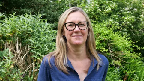 HIWWT Debbie Tann stands in front of bushes and trees. She has long, fair hair and wears glasses and a dark blue shirt.