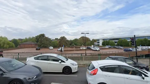 An open air car park next to a road filled with cars. 