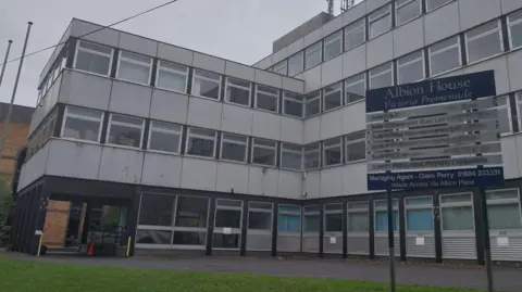 A large grey office block. The main part is four-storey, with a smaller three-storey section attached. There is a blue and silver sign out the front that says Albion House.