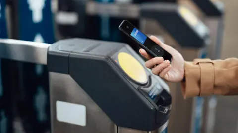 A person holding a mobile wallet up to a ticket barrier, either at a train station or a subway