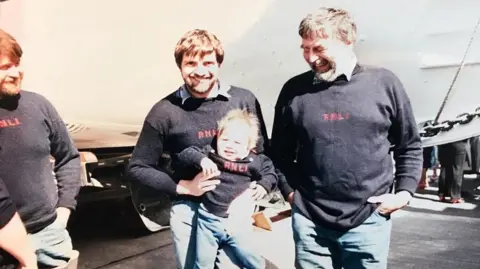 Whiteley Family An old photograph of Dave, James and Tank Whiteley all wearing blue jeans and navy blue jumpers with RNLI embroidered on them in red.