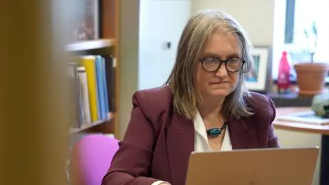 Eloise Alanna/BBC Elizabeth Elbourne is seen at her office looking at a laptop. She has straight, shoulder-length grey and brown hair, and wears round black-framed sunglasses, a burgundy suit jacket, a white blouse underneath and a necklace with a large dark green stone. Behind her is a desk and a bookshelf. 