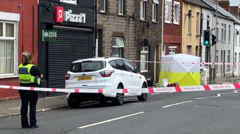 BBC A forensic white and yellow tent on Barry Road. A police officer stands watching with her back to the photographer. Police tape cordons off the area.
