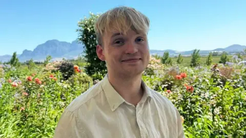 A photo of Erik Ibsen smiling at the camera. He has blonde hair that comes down to just above his eyes and is wearing a very light coloured yellow shirt that is buttoned up. Behind him is a picturesque scene of green bushes and general greenery with some mountains in the far background.