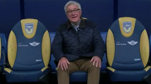 Nick Harris sitting in the blue and yellow sub-bench chairs at Oxford's Kassam Stadium.