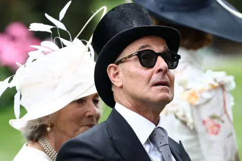 Reuters Actor Stanley Tucci looking up at something at Royal Ascot. He is wearing sunglasses and is wearing a suit.