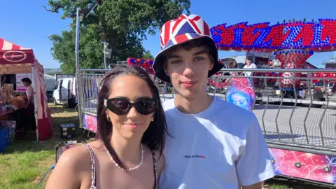 A young woman and young man are standing together. The woman has sunglasses on the the man is wearing a hat with a union flag design on it. An amusement park ride is behind them. A stall selling food is in the background.
