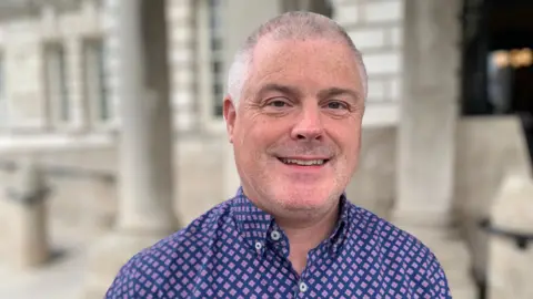 Councillor Michael Long with short grey hair is smiling at the camera. He is wearing a purple shirt with a diamond pattern on it. 