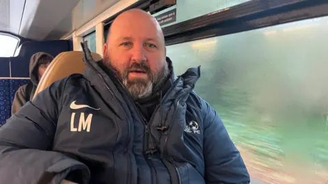 Leon Mumford on board a bus. He is sat next to a window and is wearing a blue coat. 