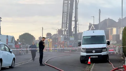 A police officer stood in the middle of the road in front of a cordon where fire engines are in place dealing with a fire. the scene is still verry smoky.