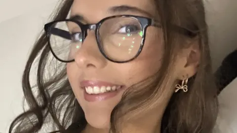 PA Media A close-up picture of Amber looking at the camera. She has long dark hair and dark-rimmed glasses.