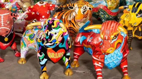 Close-up photos of a group of nearly life-size fibre-glass statues of bulls, brightly painted in various designs. The one in the foreground at the front of the picture has a red heart painted on its chest with the letters BRUM in gold inside. Another next to it is painted red and blue to look like a chinese Dragon.