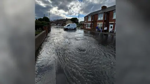 The road and the left-side pavement of Ellen Terrace covered in water, which flows down the street. A group of five people is standing outside one of the terraced houses and looking on. A white Northumbrian Water van is parked horizontally at the top of the street.