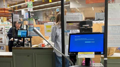 A blue error screen can be seen on a checkout till at a supermarket in Australia. It is in the foreground, and behind it, a woman can be seen paying at another till.