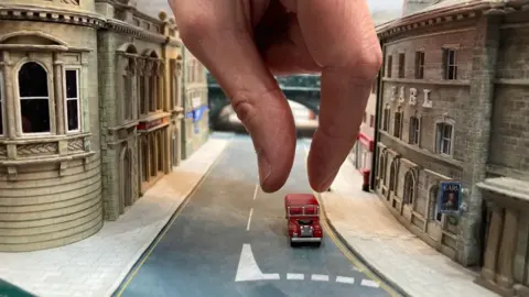 A human hand reaching down into a model street scene to pick up a red minature model of a landrover