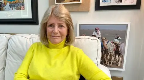 BBC Linda Jackson is sitting on a sofa with framed pictures behind on her a wall. She is wearing a polo neck yellow jumper and has blonde hair.
