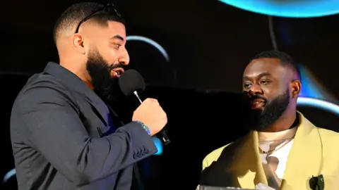 Getty Images James Duncan and Fuhad Dawodu receiving a Mobo Award in February. James has short hair and a black beard, he wears sunglasses on his head and a blue suit, holding a mic. He's looking at Fuhad, who also has short hair and a black beard and is wearing a greeny-yellow suit over a white top. The staging behind them is black with streaks of blue.  