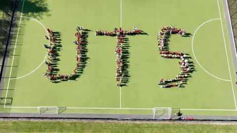 Moygashel Regeneration Group An overhead shot of a green football pitch with white markings. On the pitch dozens of children are standing in formation which spells out UTS, for Up The Swifts. 