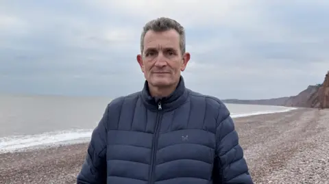 Mike Brown stood on the beach. He is looking at the camera and is wearing a blue coat.